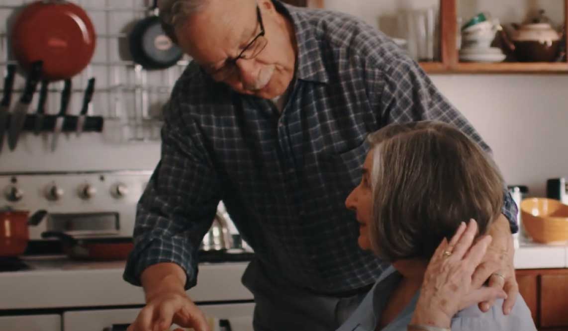 An elderly couple hugging in their kitchen.